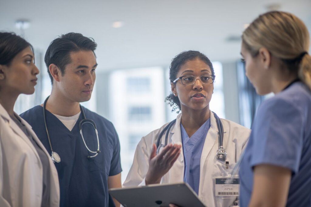 A small group of medial professionals stand together in the hallway while briefly meeting to go over a patient file.   The female doctor in a white lab coat is leading the meeting while holding out a tablet.  Her colleagues are listening attentively as she goes over the patients plan of care.